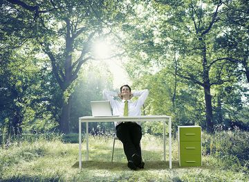 Geschäftsmann im Anzug sitzt entspannt an einem Schreibtisch, der mitten im grünen Wald steht 